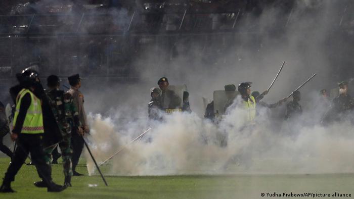Aparat kepolisian bersenjata pentungan berdiri di antara kepulan gas air mata di taman Stadion Kanjuruhan